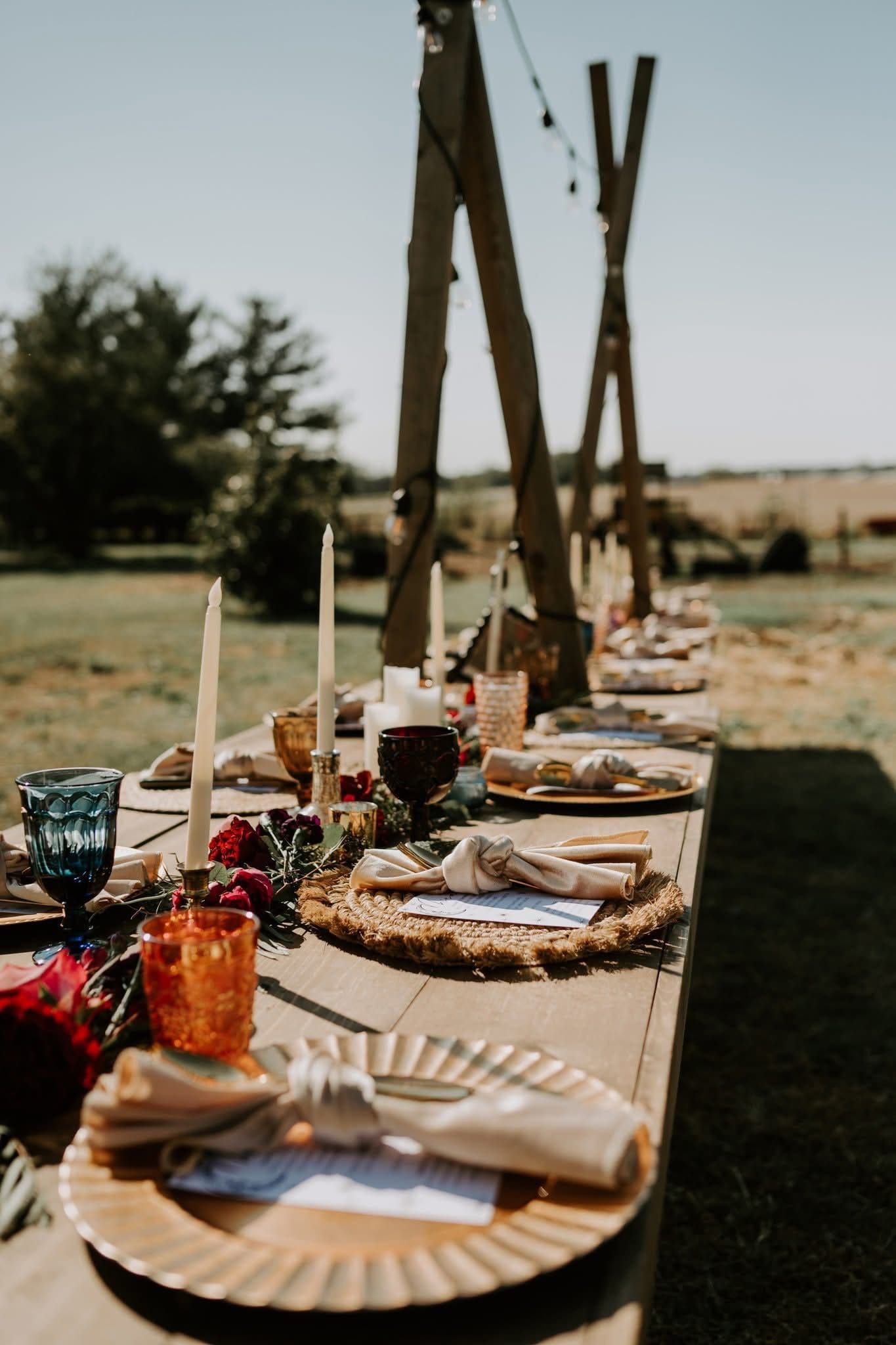 A long outdoor wooden banquet table under a timber arbor in a sunlit field, set with taper candles, rattan chargers, knotted linen napkins, coloured goblets and a deep red floral garland.