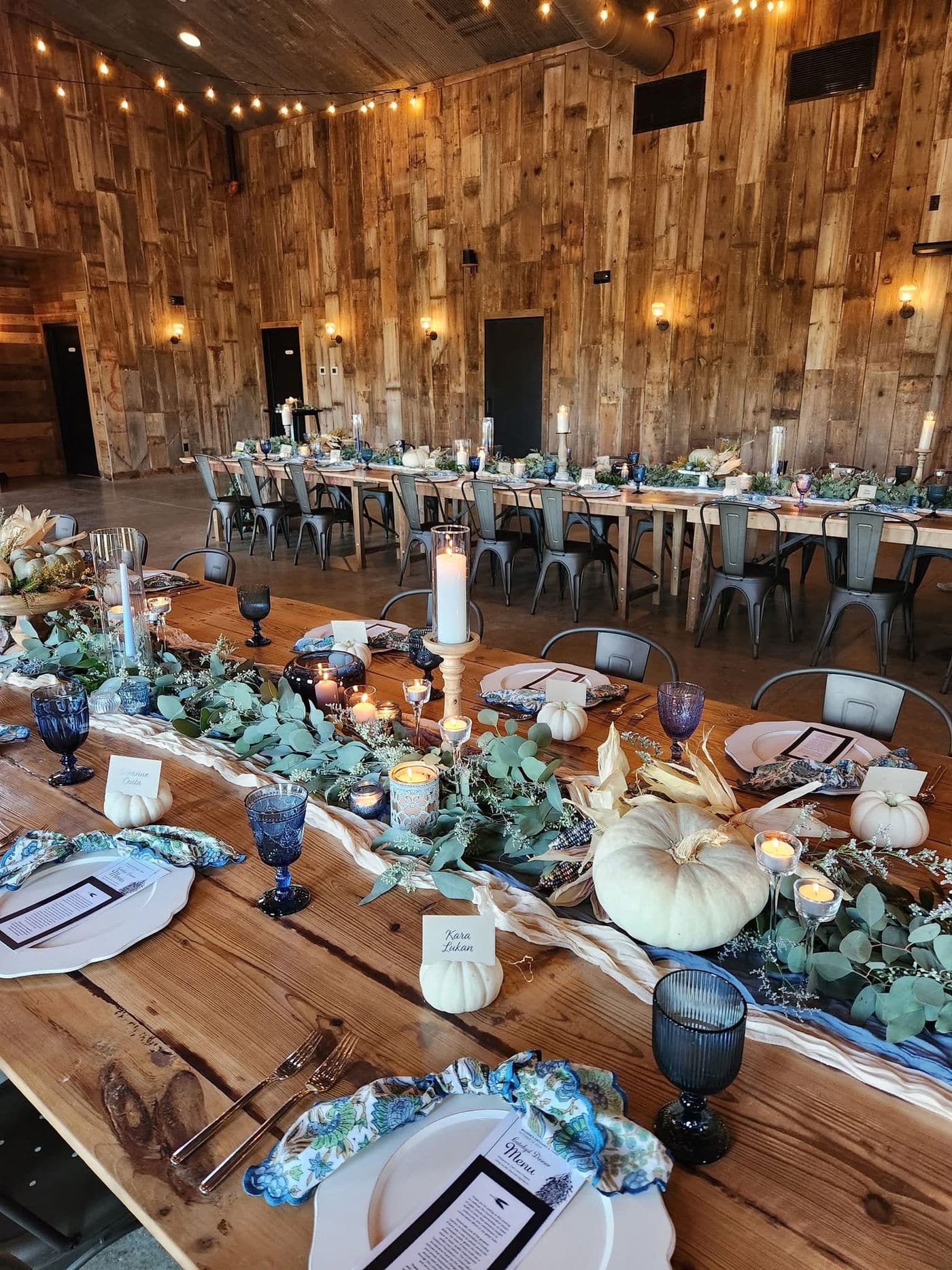 Long farmhouse tables in a rustic wood barn set for an autumn wedding reception with cobalt blue goblets, white pumpkins, eucalyptus garland, pillar candles and string lights.