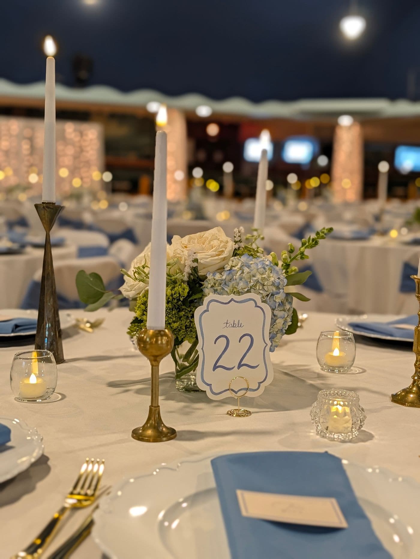A candlelit wedding reception table set with white roses and blue hydrangeas, gold taper candleholders, dusty blue napkins and a hand lettered table card in a warmly lit ballroom.