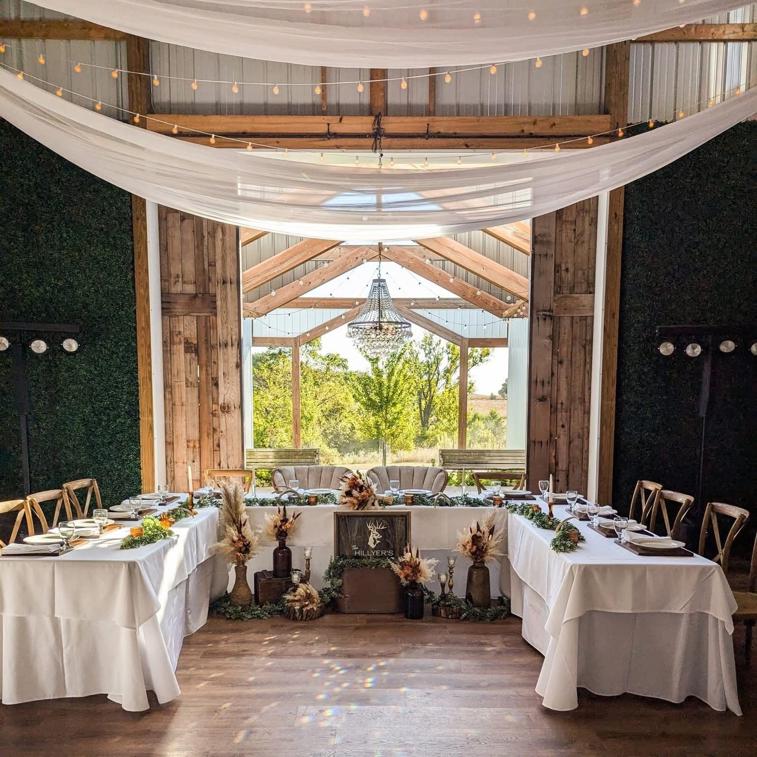 A barn wedding venue set for a reception, with white draped head tables, pampas and eucalyptus arrangements, a crystal chandelier, string lights and greenery walls framing sunlit trees.