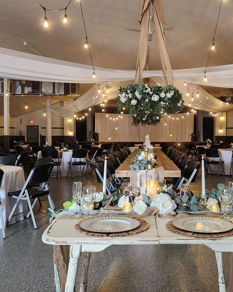 A plain community hall transformed for a wedding reception, with a distressed sweetheart table in the foreground, ceiling draping, string lights and a suspended floral wreath.