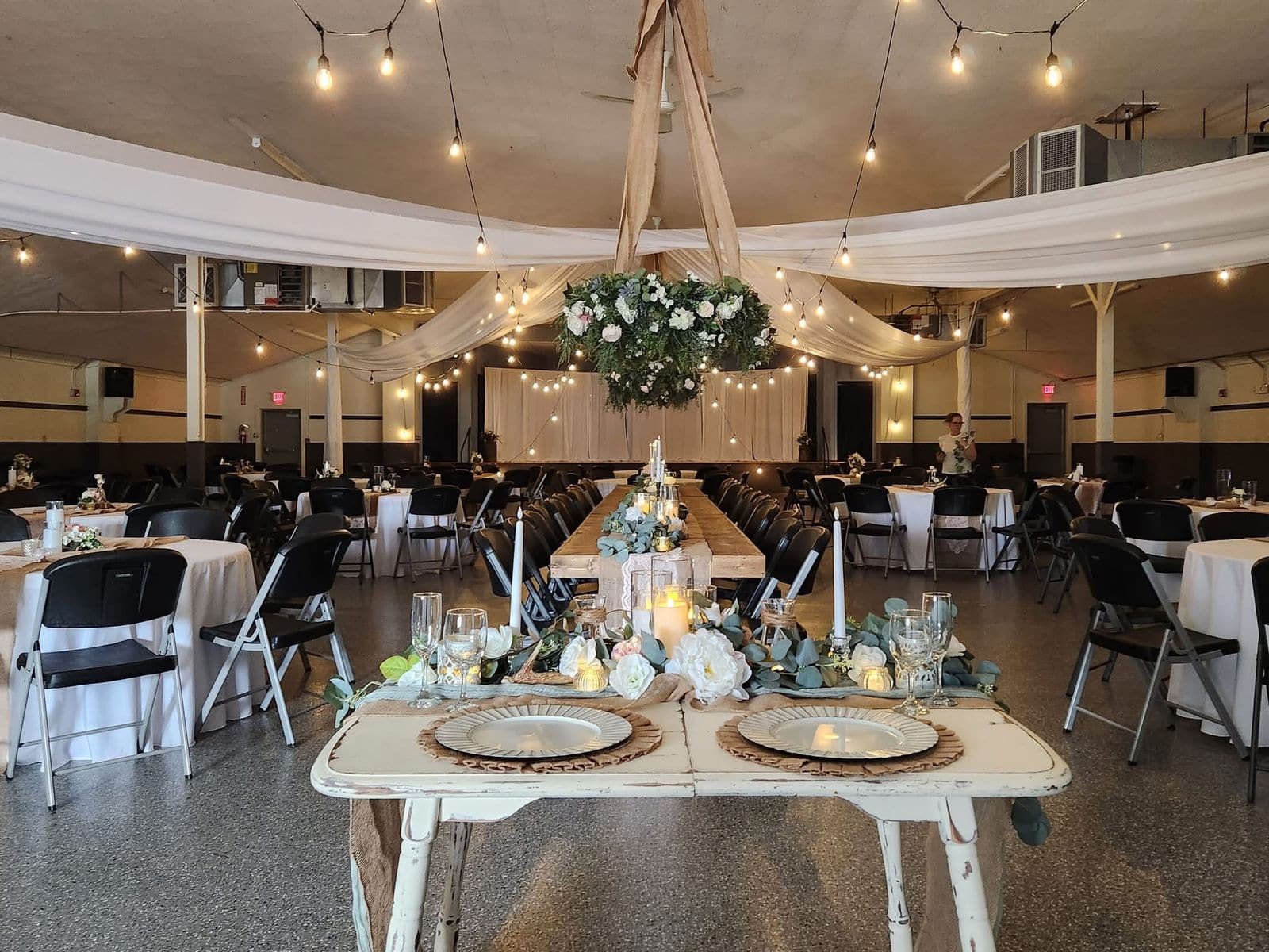 A plain community hall transformed for a wedding reception, with a distressed sweetheart table in the foreground, ceiling draping, string lights and a suspended floral wreath.