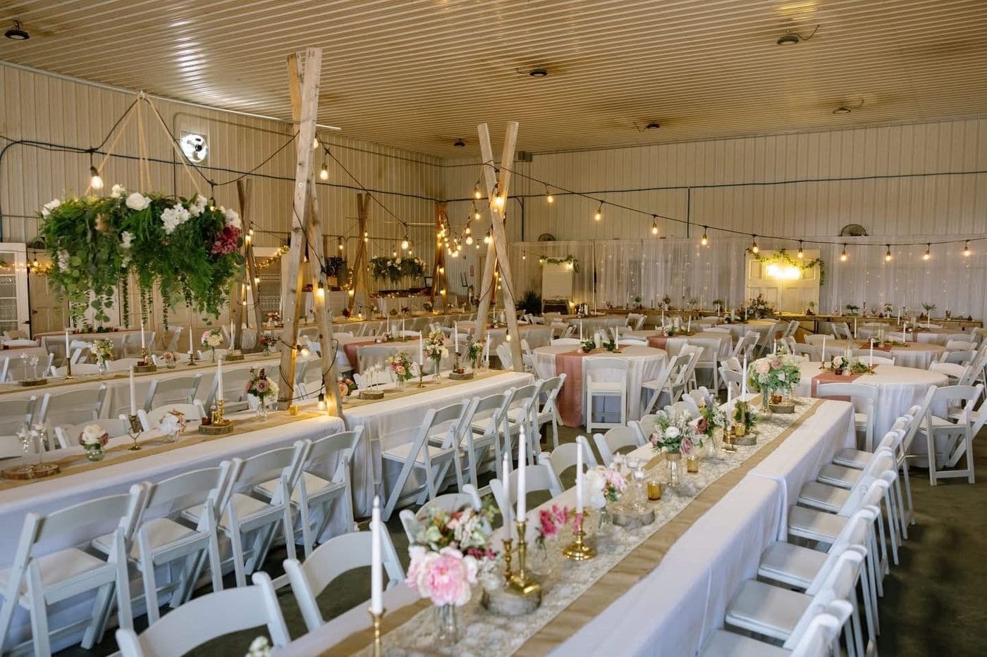 A rustic indoor wedding reception hall set with long white linen tables, blush and burlap runners, white folding chairs, gold candlesticks, string lights and a suspended greenery chandelier.