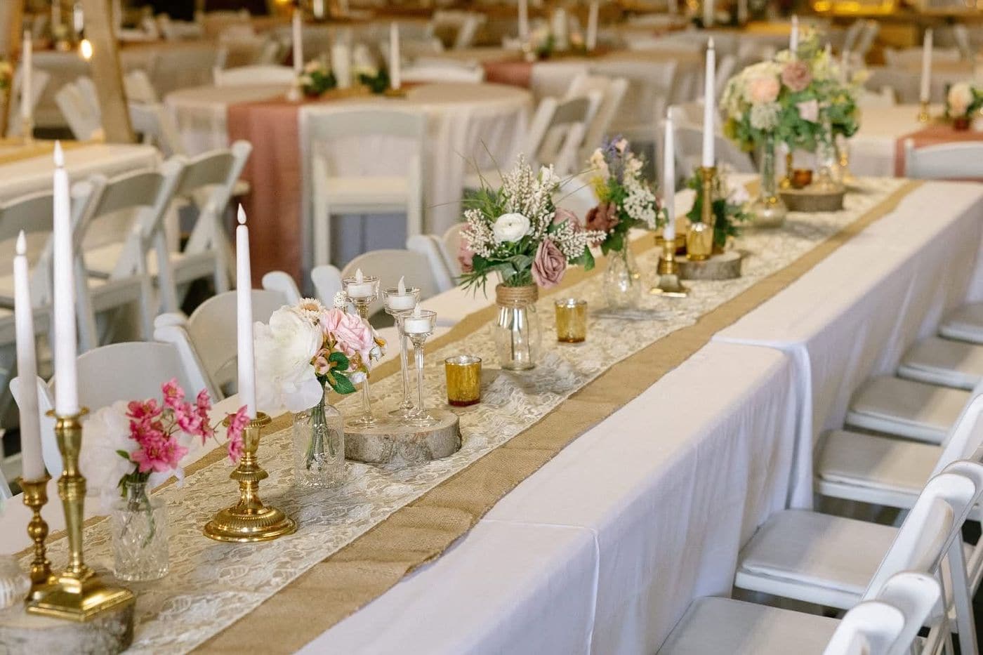 A long rustic wedding table set with a burlap and lace runner, brass candlesticks, mason jar and bud vase florals in pink and cream, and wood slice risers.