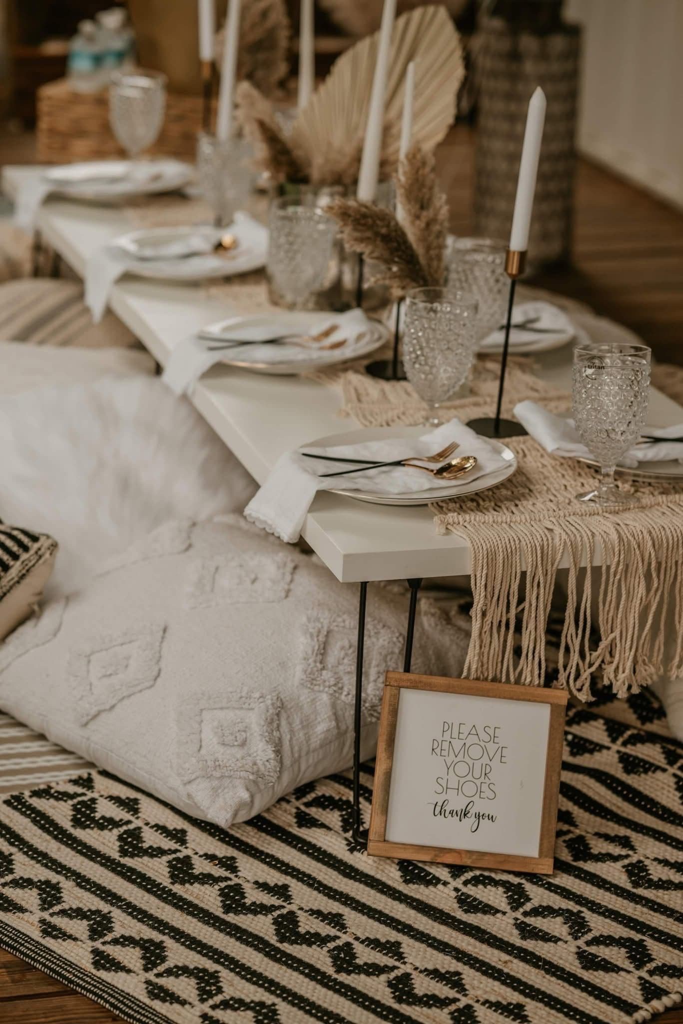 A low boho picnic table set with white plates, gold flatware, taper candles, pampas grass, a macrame runner and floor cushions on a patterned jute rug.