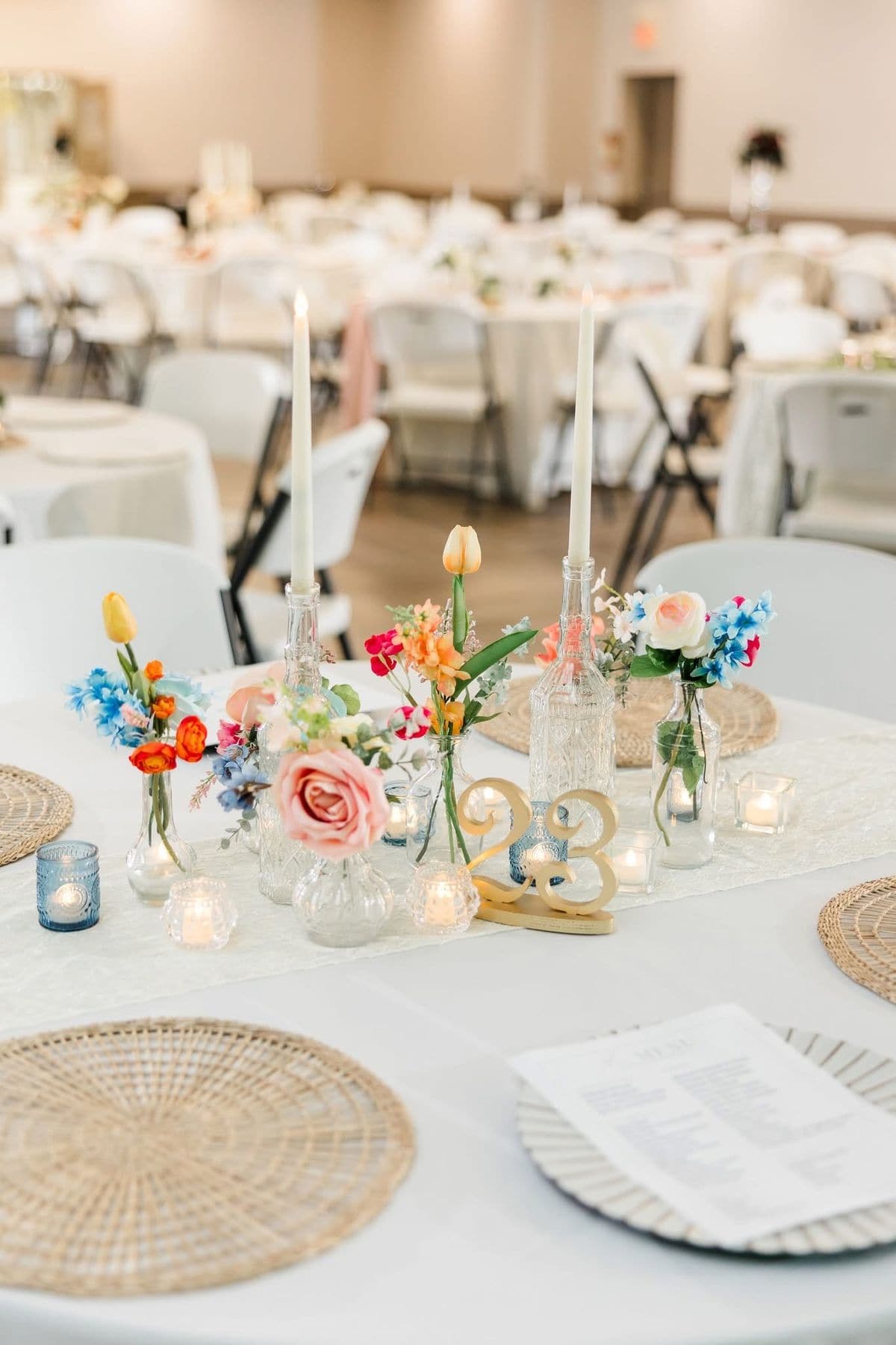 A colourful bud vase centrepiece of tulips, roses and blue blooms with taper candles in vintage glass bottles, a gold table number, rattan placemats and a printed menu.