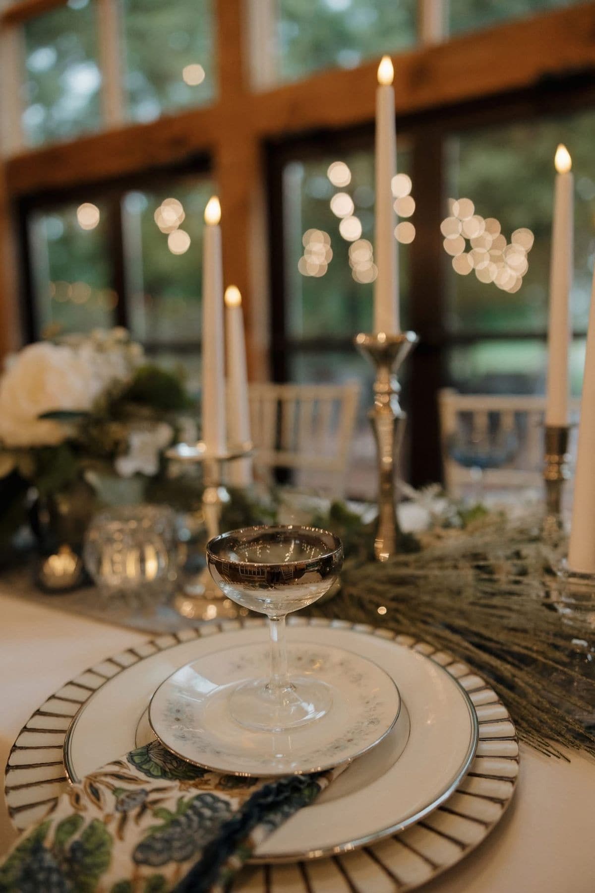 A candlelit wedding place setting, a silver rimmed coupe glass on layered vintage china with a floral napkin, greenery runner and taper candles glowing against tall windows.