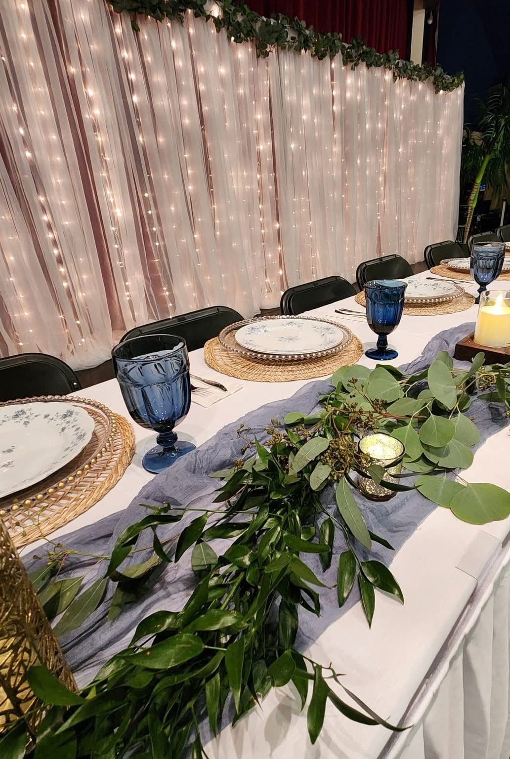 A banquet table set with cobalt blue goblets, rattan chargers, floral china, a dusty blue gauze runner and trailing eucalyptus, backed by a blush curtain strung with fairy lights.