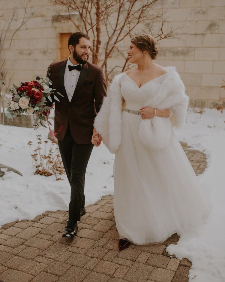 A newlywed couple walk hand in hand along a brick path in the snow, the bride in a white gown and fur stole, the groom in a burgundy tuxedo holding a deep red bouquet.