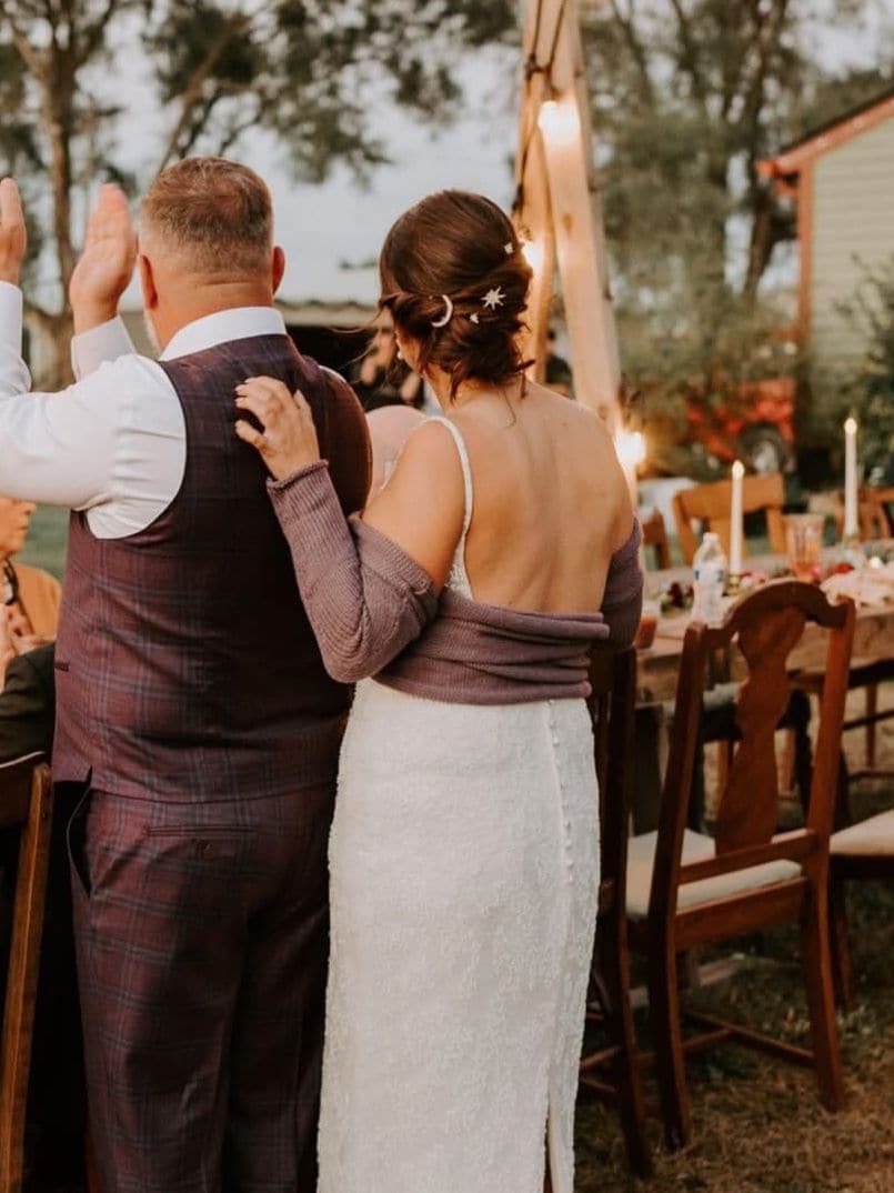 A newly married couple stand arm in arm with their backs to the camera, toasting their guests at an outdoor reception at dusk under string lights.