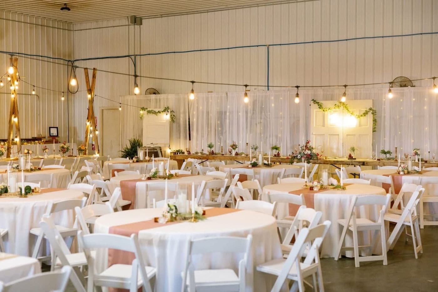 A barn style venue set for a wedding reception with white linen round tables, blush runners, white folding chairs, string lights and a sheer draped backdrop with fairy lights.