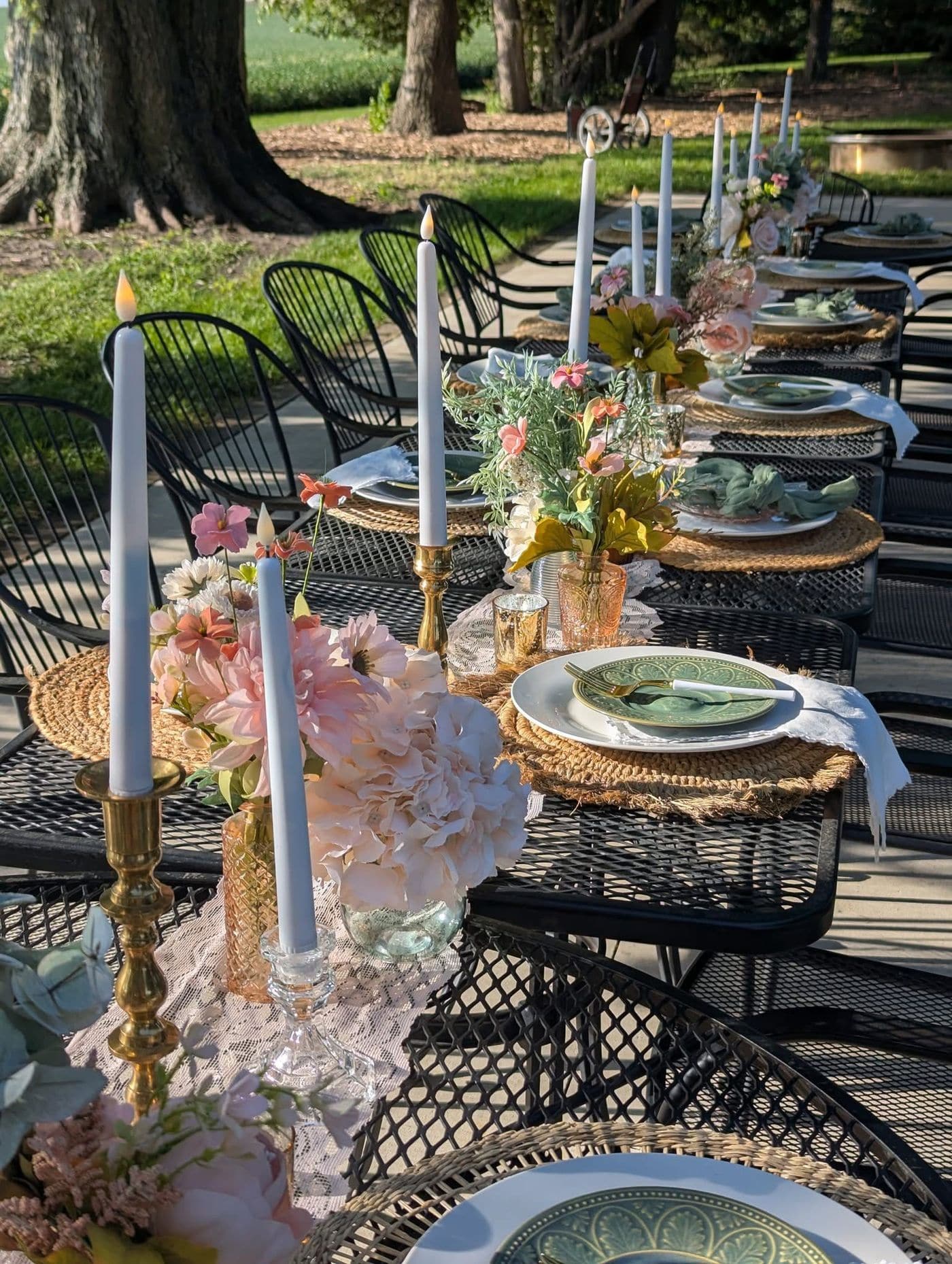 A long outdoor table under shade trees with black wrought iron furniture, rattan placemats, green scalloped plates, brass candlesticks and blush floral arrangements in amber glass vases.
