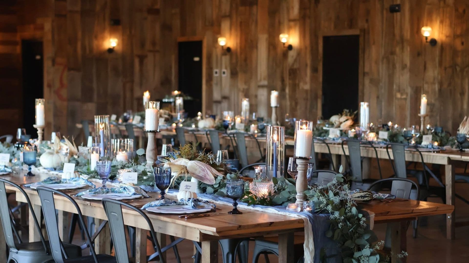 Long farmhouse banquet tables in a warm wood barn set for a wedding reception, with cobalt blue goblets, pillar candles in glass hurricanes, eucalyptus garland and dusty blue runners.