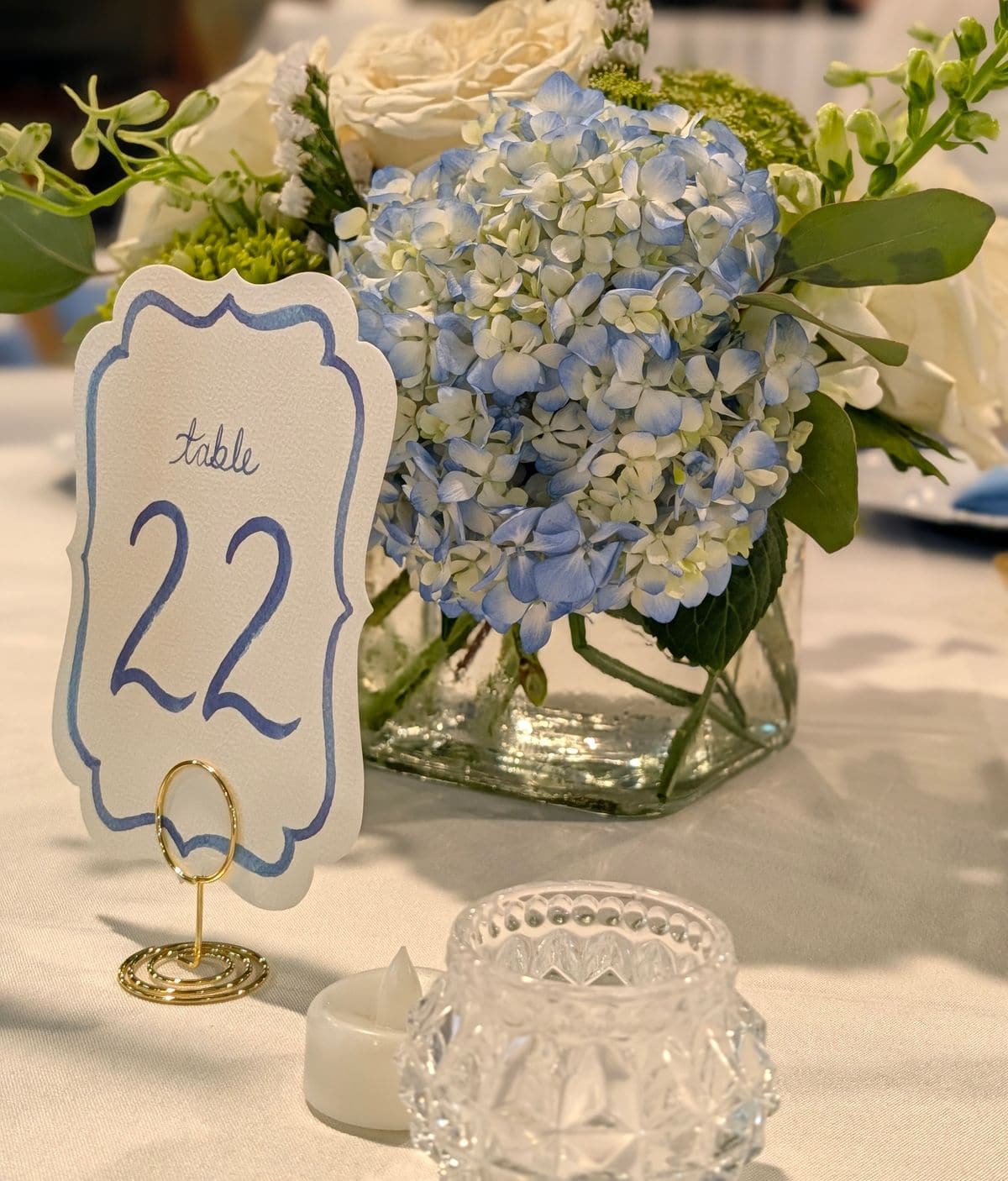 A close detail of a wedding centrepiece of blue hydrangeas and white roses in a square glass vase, beside a hand lettered table card and a cut glass votive holder on white linen.