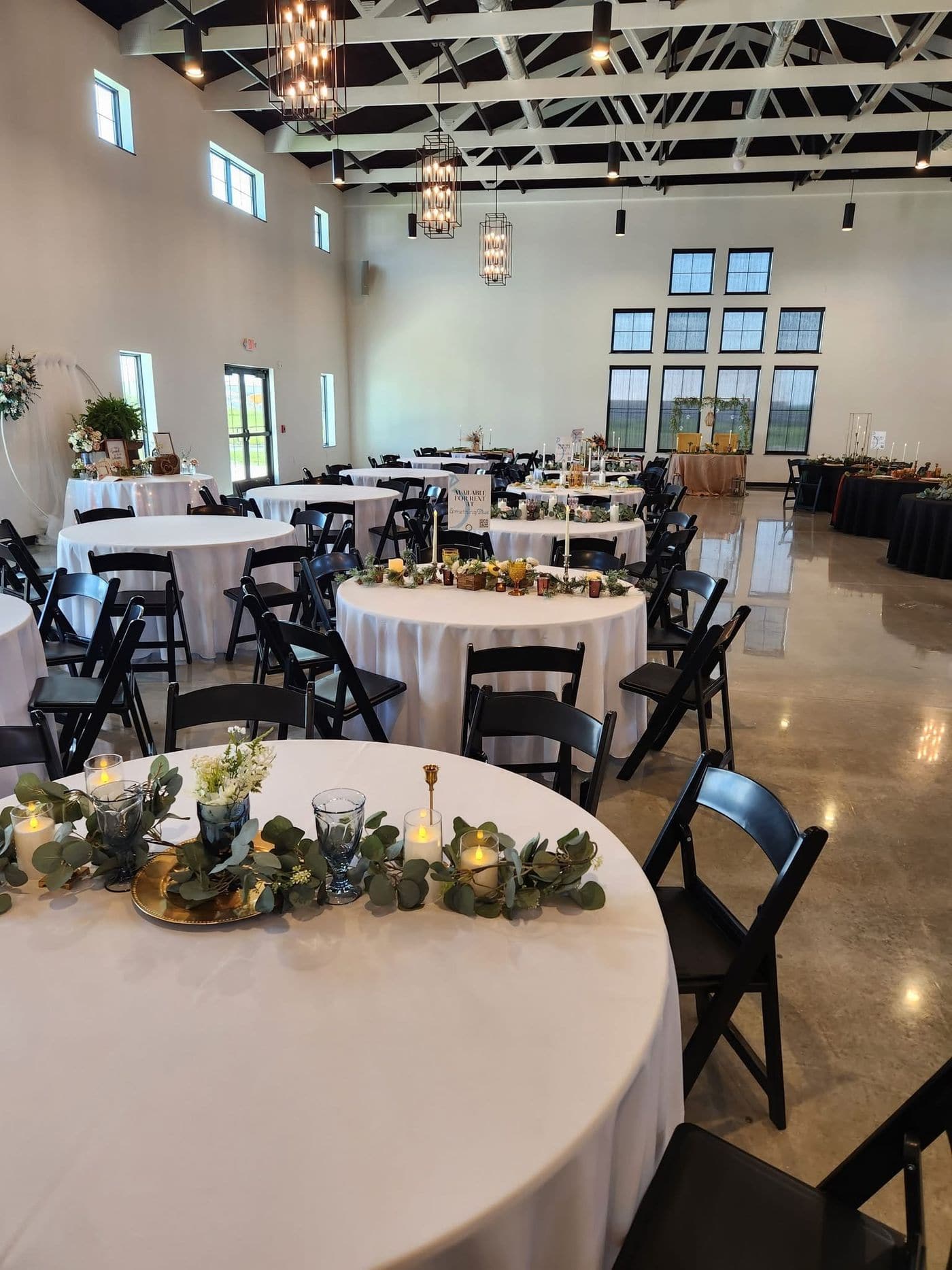 A spacious event hall set for a wedding reception with round white linen tables, black folding chairs, eucalyptus and candle centrepieces, black chandeliers and tall windows over open fields.