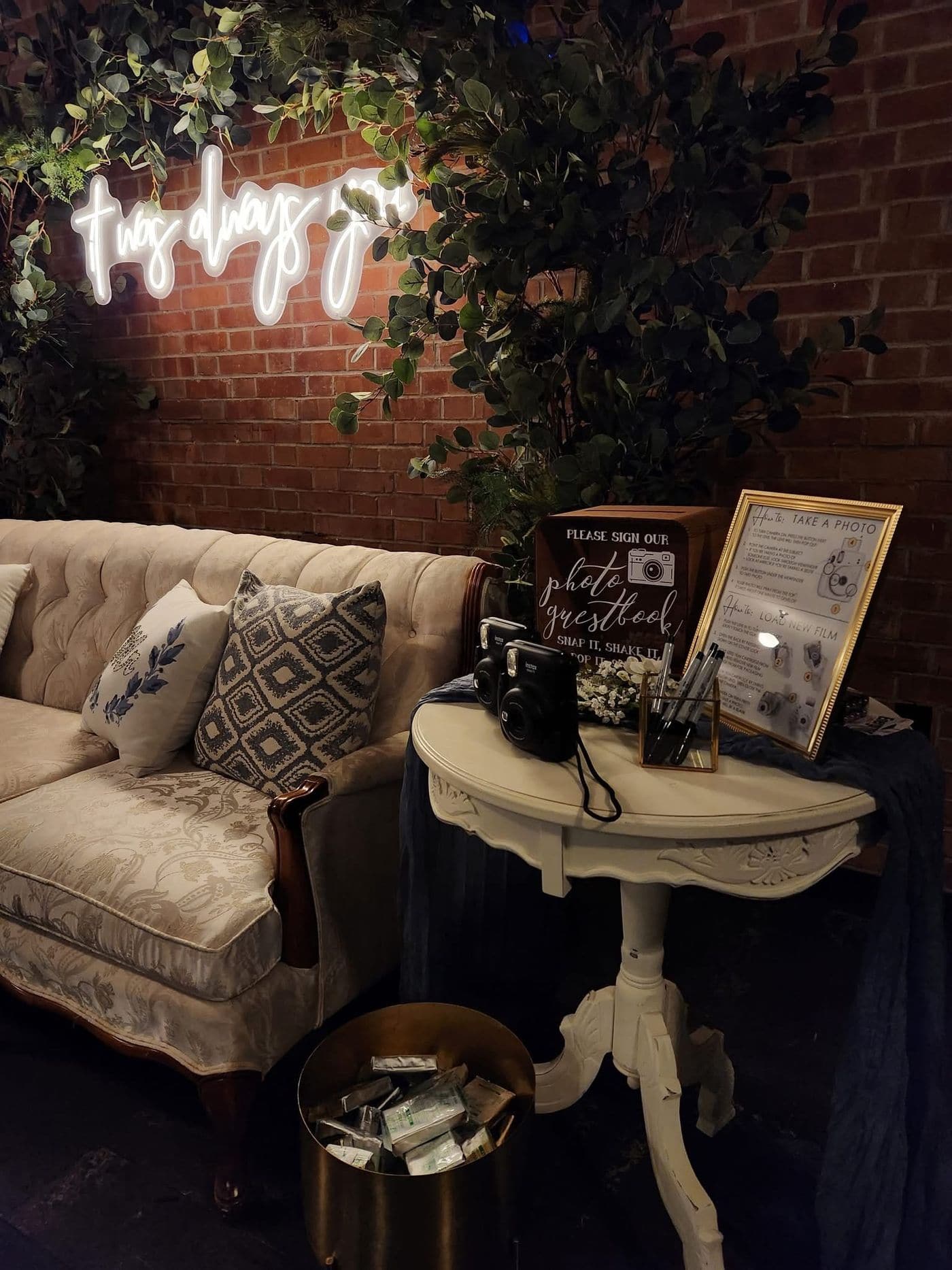 A dimly lit reception lounge with a tufted ivory settee, a pedestal table set up as an instant photo guestbook, eucalyptus over exposed brick and a glowing neon sign reading twas always you.