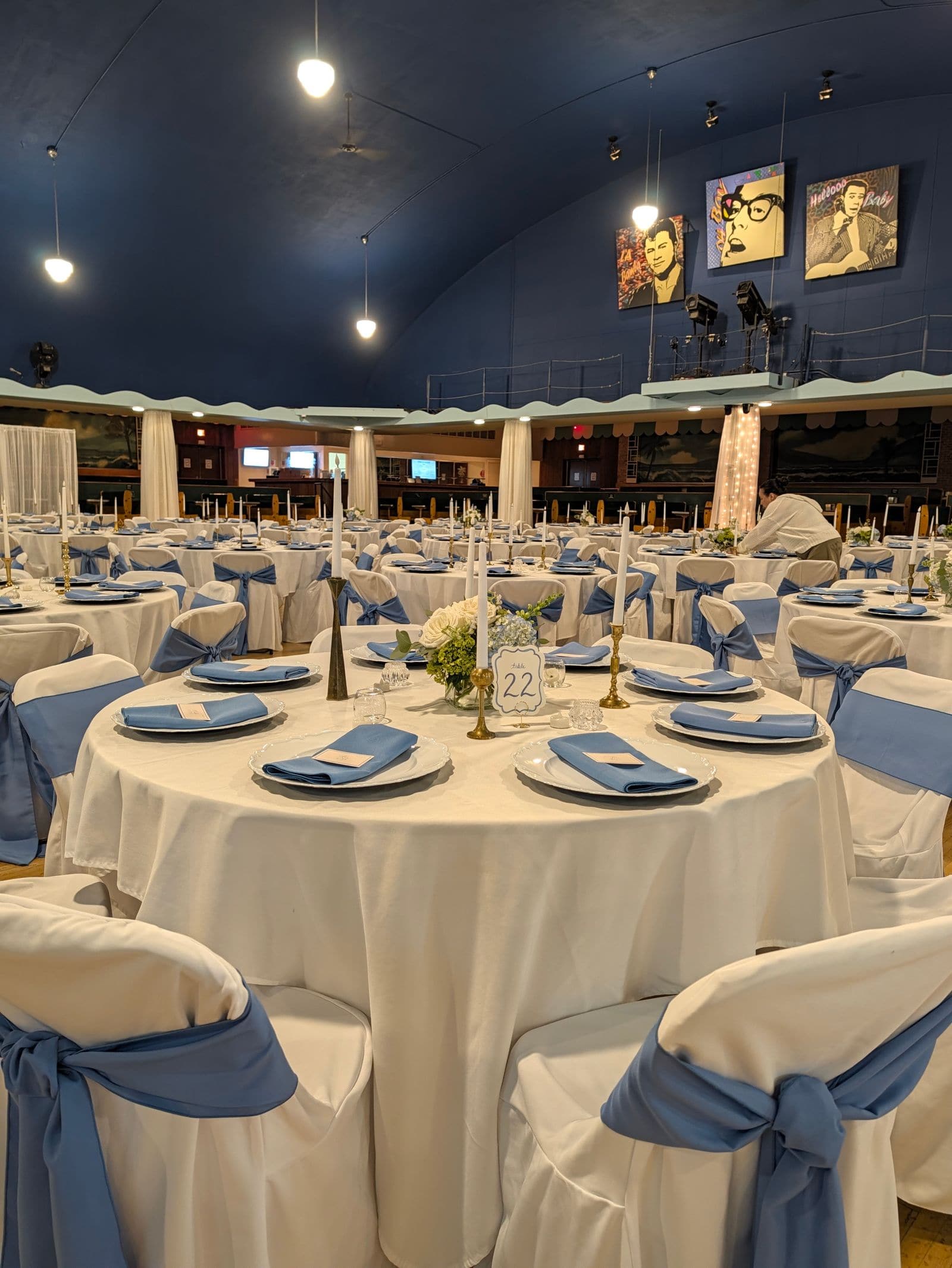 A full wedding reception hall under a vaulted navy ceiling, with round tables in white linen, dusty blue chair sashes and napkins, white rose and hydrangea centrepieces and taper candles.