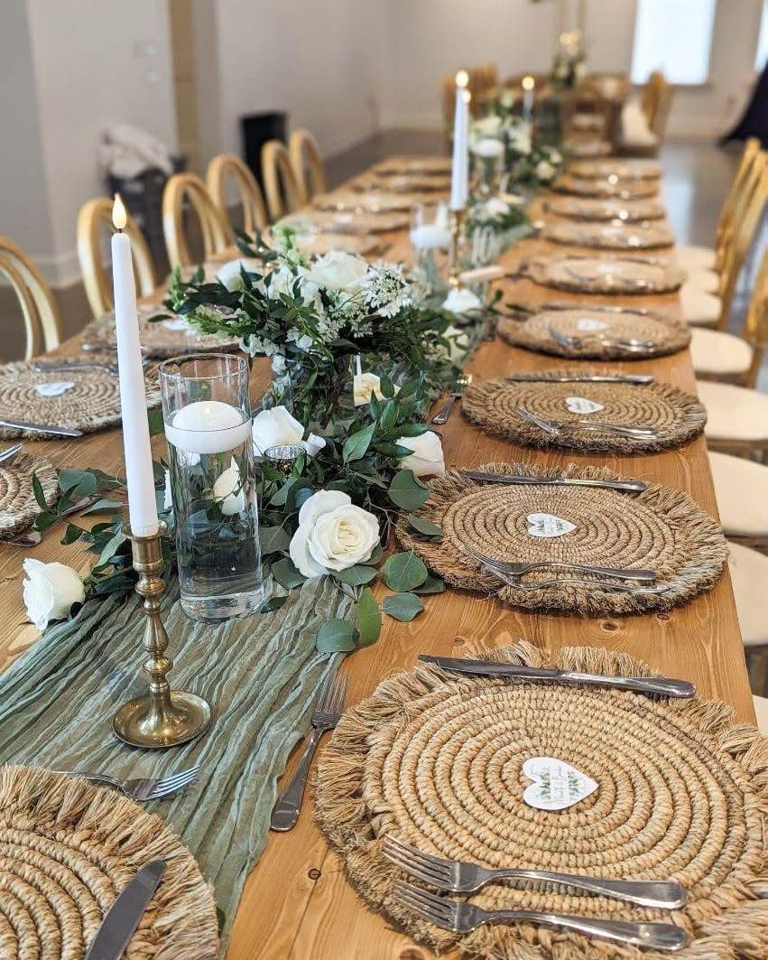 A long wooden table set with jute chargers, brass candlesticks, floating candles, a sage gauze runner and white rose and eucalyptus centrepieces.