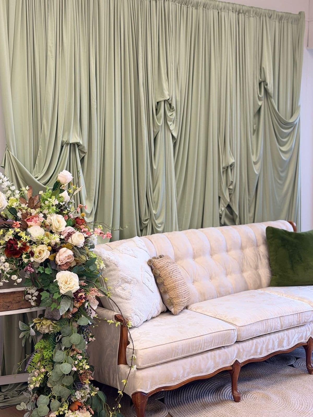 A vintage tufted cream settee styled with textured pillows and a cascading floral arrangement against a sage green draped backdrop.