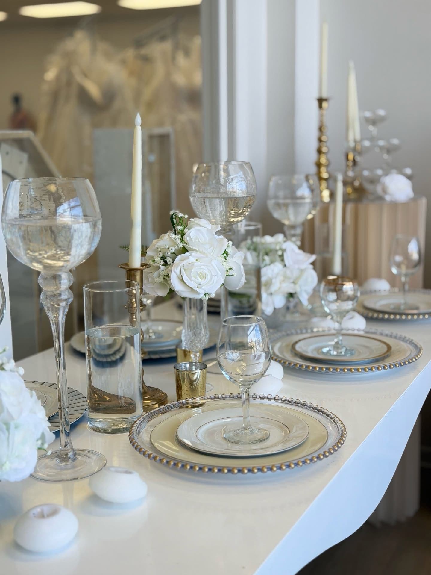 A white and gold wedding tablescape with beaded chargers, crystal stemware, taper candles and white roses in the Something Blue showroom.