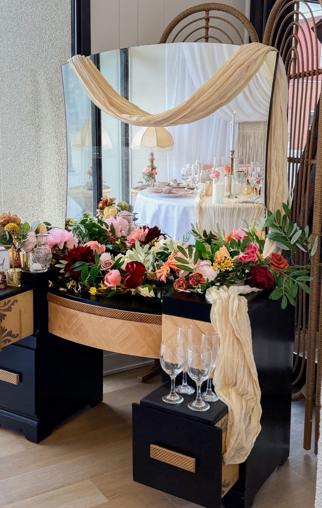A refinished black and wood vanity with an arched mirror, styled as a wedding welcome display with a floral garland and champagne flutes.