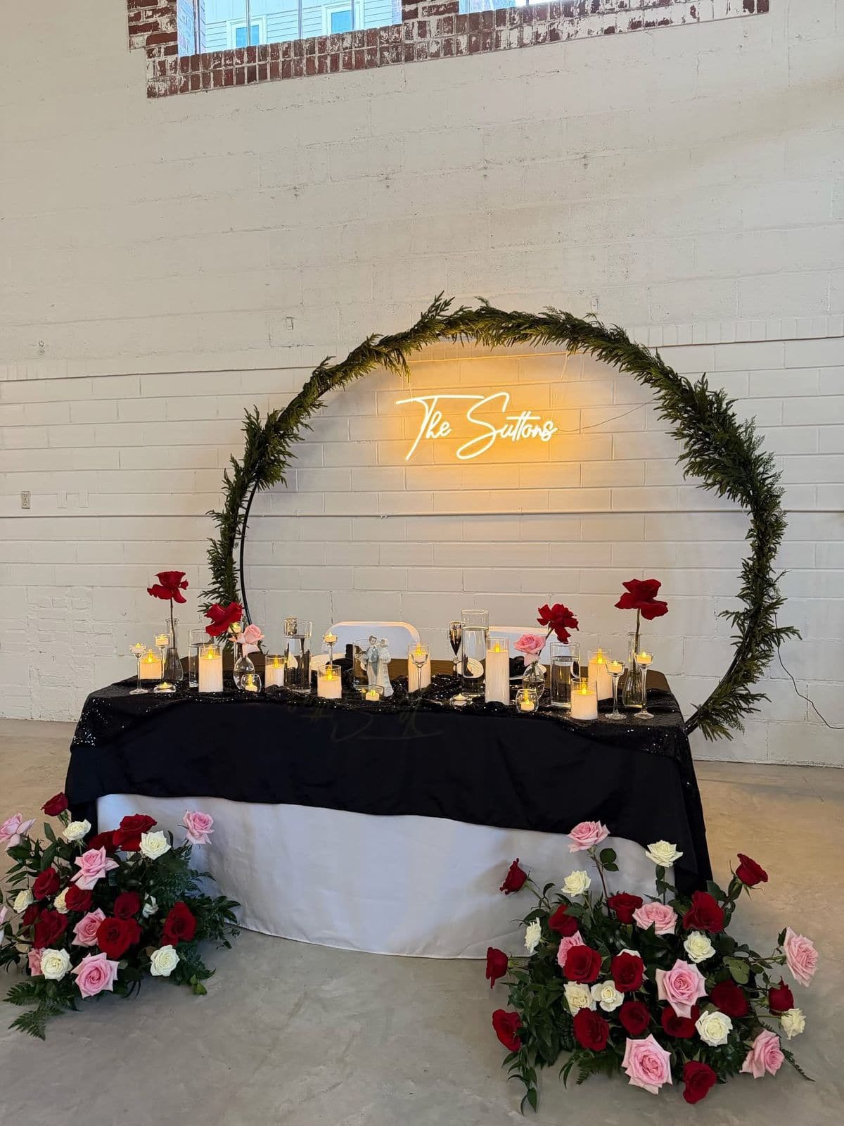A sweetheart table in black and white linen with a greenery wrapped circular arch framing a glowing neon sign, flanked by red, pink and white rose arrangements against white brick.