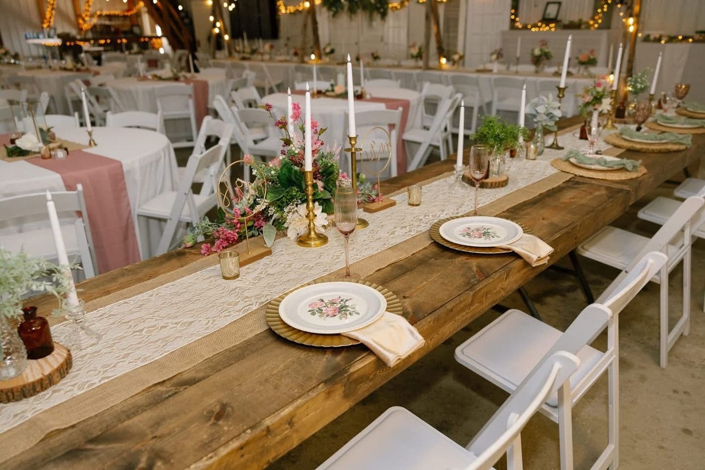 A reclaimed wood farm table set with a lace runner, gold chargers, floral vintage china, brass taper holders and pink garden flower centrepieces, surrounded by white folding chairs.