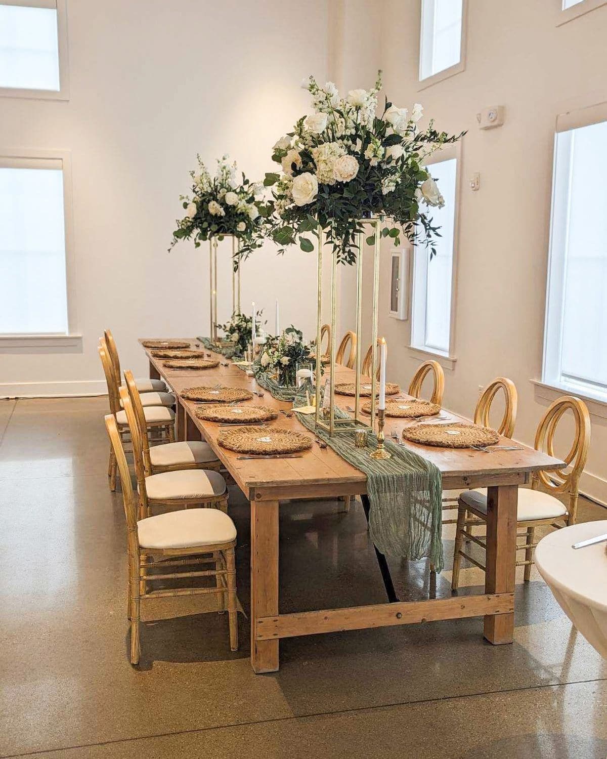 A long farmhouse table in a bright white venue with tall white and greenery pedestal arrangements, a sage gauze runner, rattan placemats and cane back chairs.
